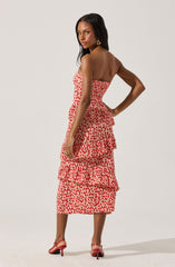 A woman is wearing a strapless, knee-length dress with a beige background and red, pink, and orange polka dot pattern. The dress features a fitted bodice and three layered ruffles on the skirt. She is also wearing pink high-heeled sandals with ankle straps and large gold hoop earrings.