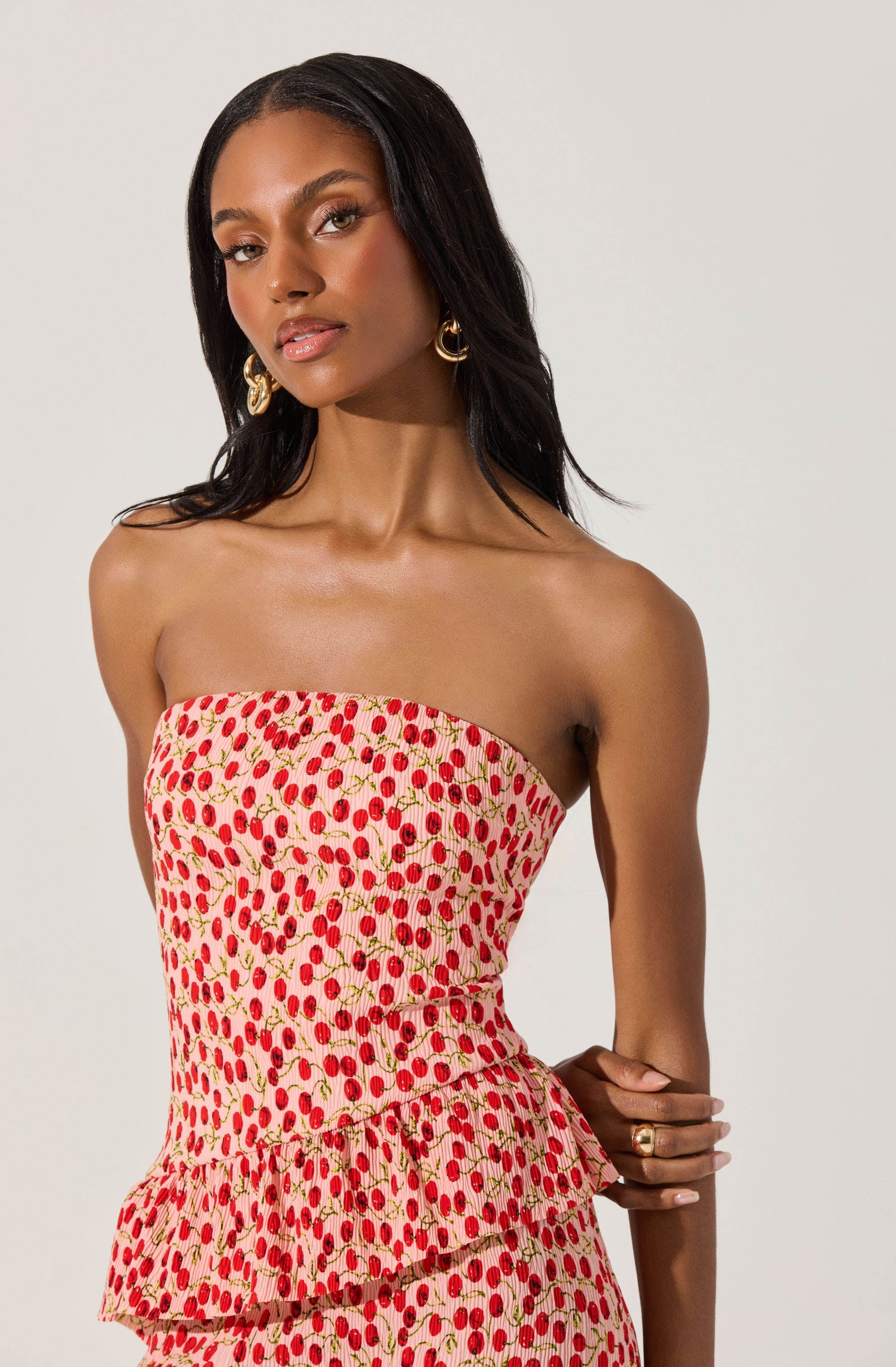 A woman is wearing a strapless dress with a fitted bodice and a peplum detail at the waist. The dress features a pattern of small red cherries with green stems on a light pink background. She accessorizes with large gold hoop earrings and a gold ring on her right hand.