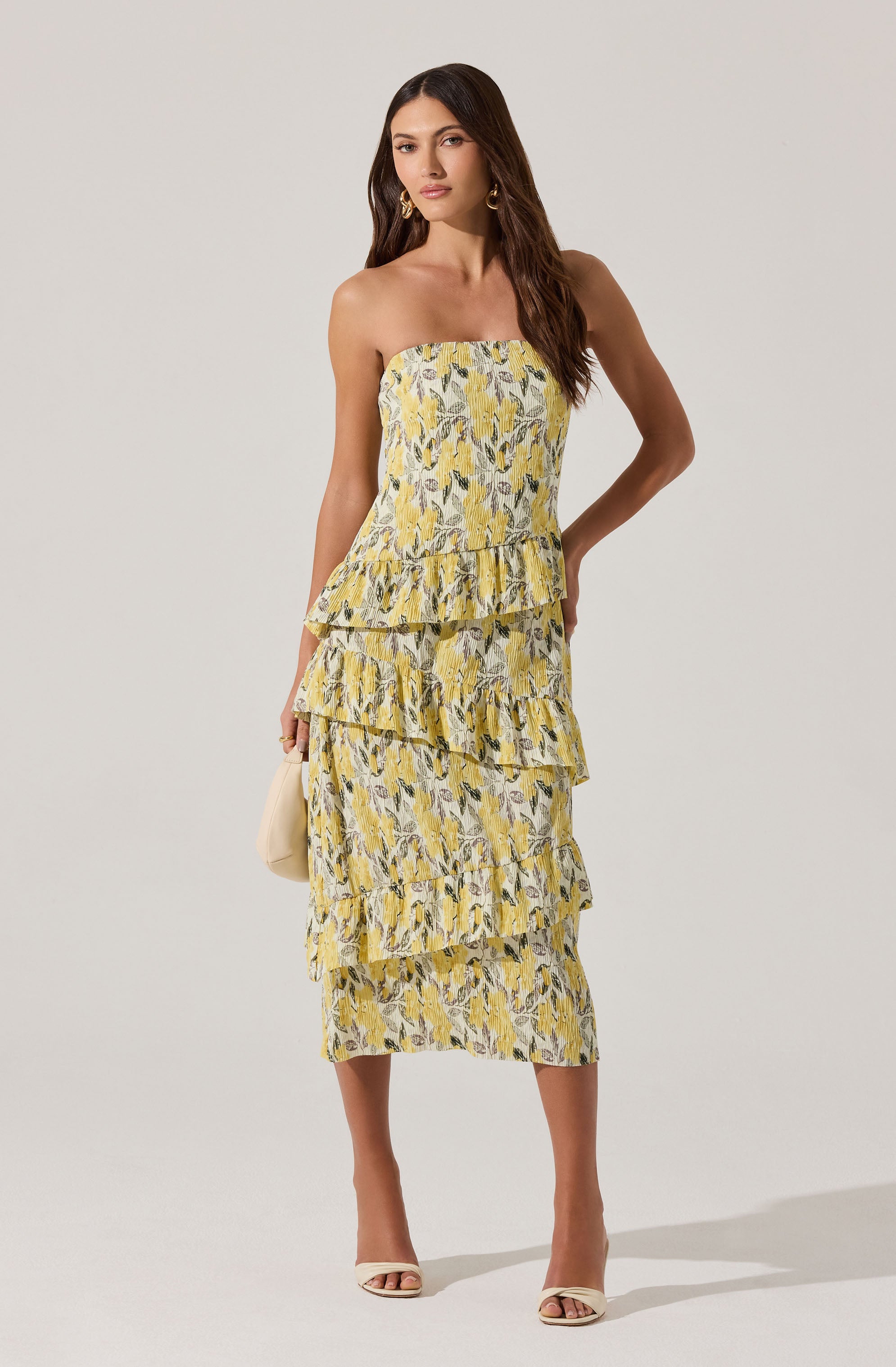 A woman is wearing a strapless, yellow floral dress with a layered ruffle design. The dress features a pattern of leaves and flowers in shades of yellow, white, and gray. She is accessorized with large gold hoop earrings and is holding a small beige handbag. She is also wearing cream-colored open-toe heels.