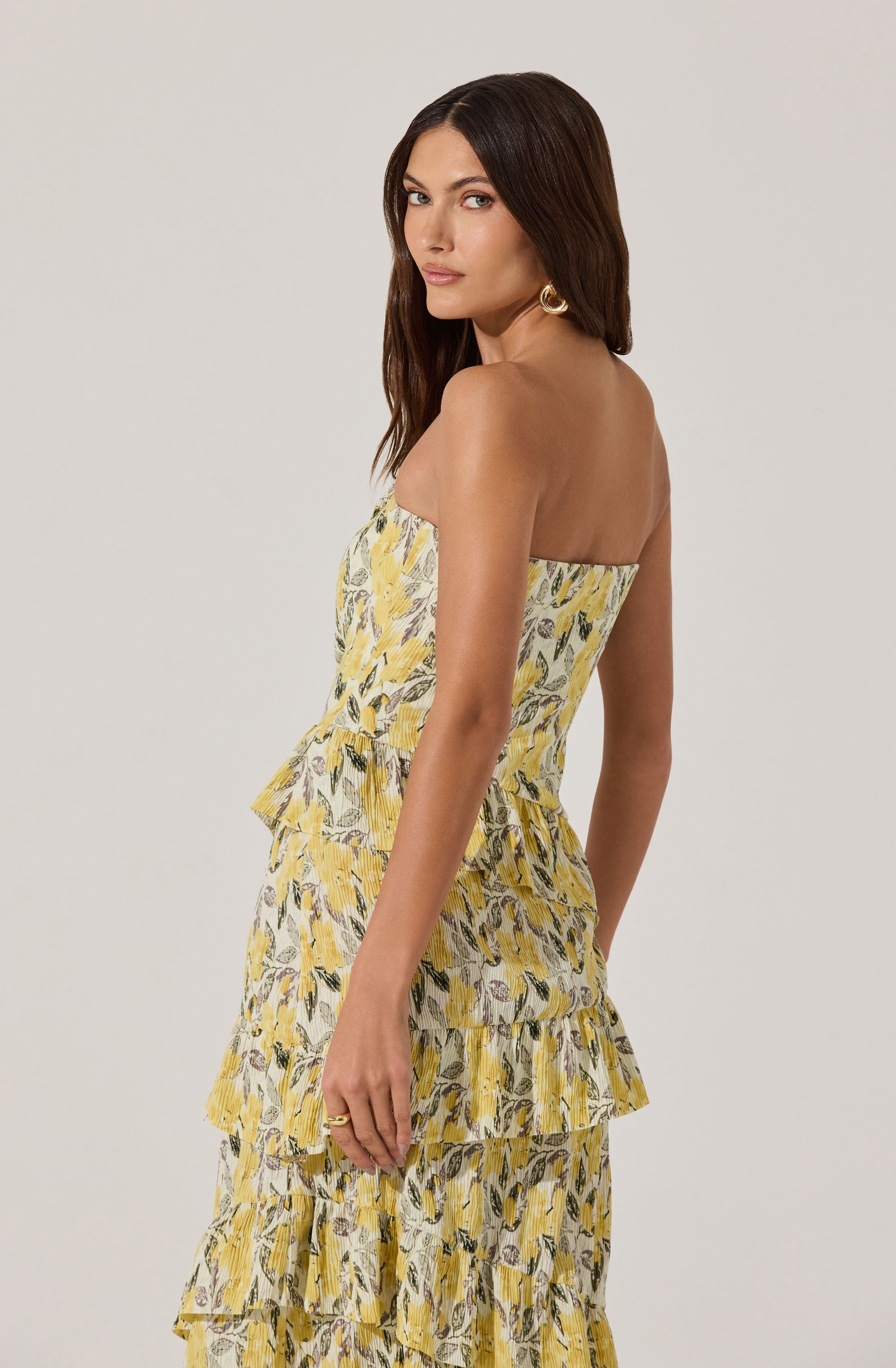 A woman is wearing a strapless, tiered maxi dress with a yellow, white, and black floral pattern. The dress features multiple ruffled layers, creating a flowing and textured appearance. She accessorizes with large gold hoop earrings.