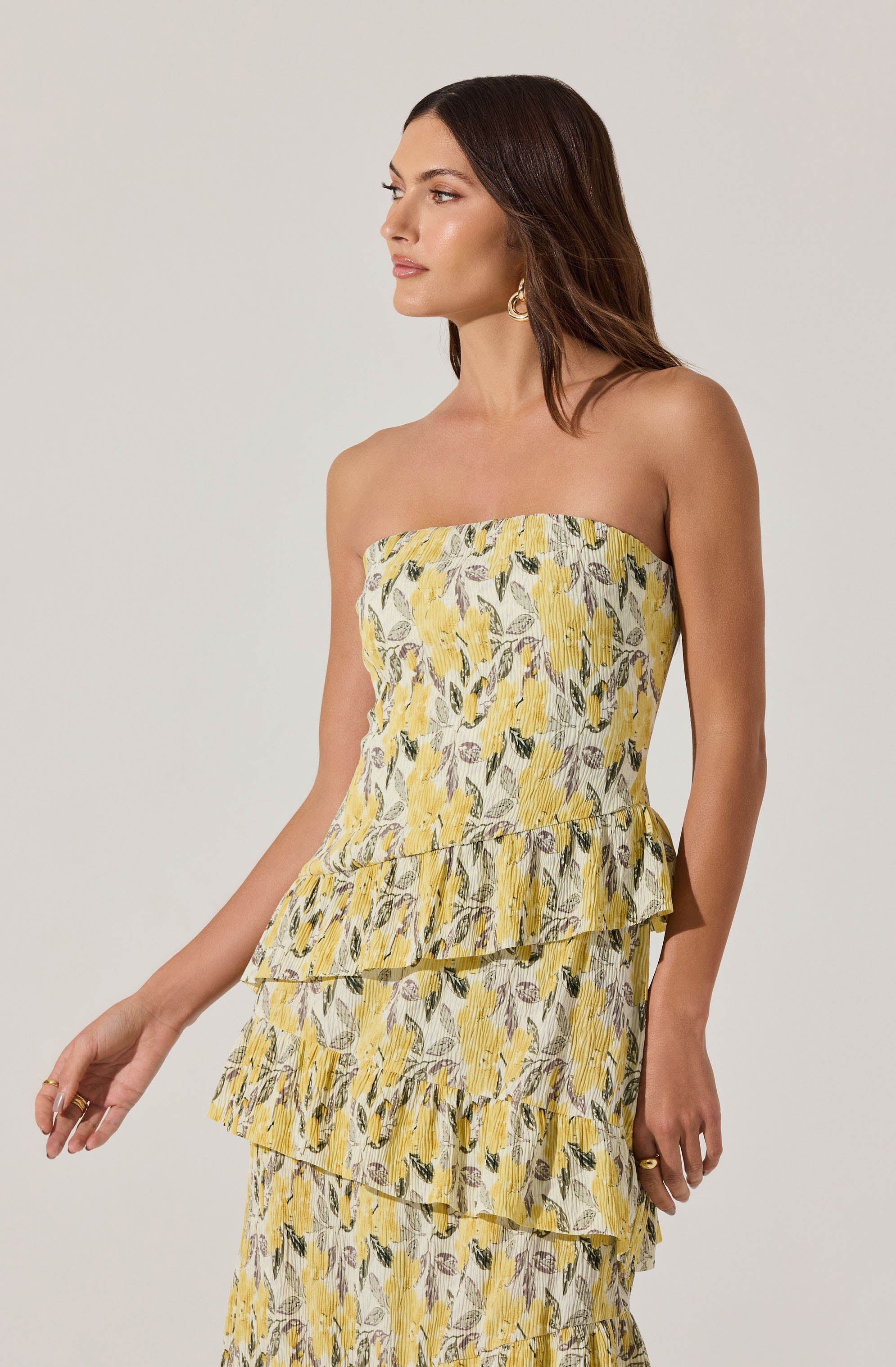 A woman is wearing a strapless dress with a fitted bodice and a tiered skirt. The dress features a yellow floral pattern with green and gray leaves on a light background. The fabric appears lightweight and slightly textured, suitable for warm weather.