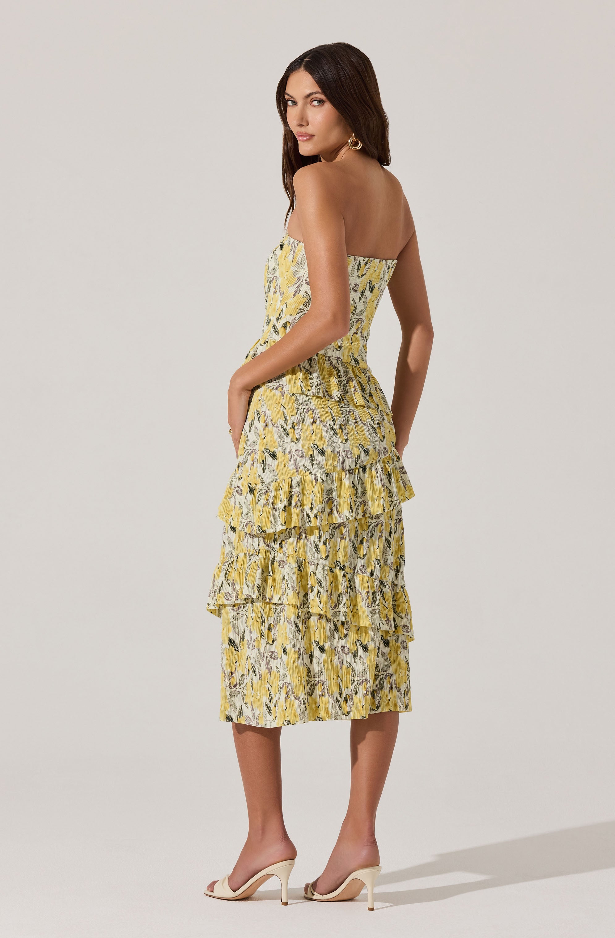 A woman is wearing a strapless, yellow and white tiered dress with a floral or abstract pattern. The dress has multiple ruffled layers that extend down to mid-calf length. She is also wearing light-colored, open-toe high-heeled shoes and large, circular earrings.
