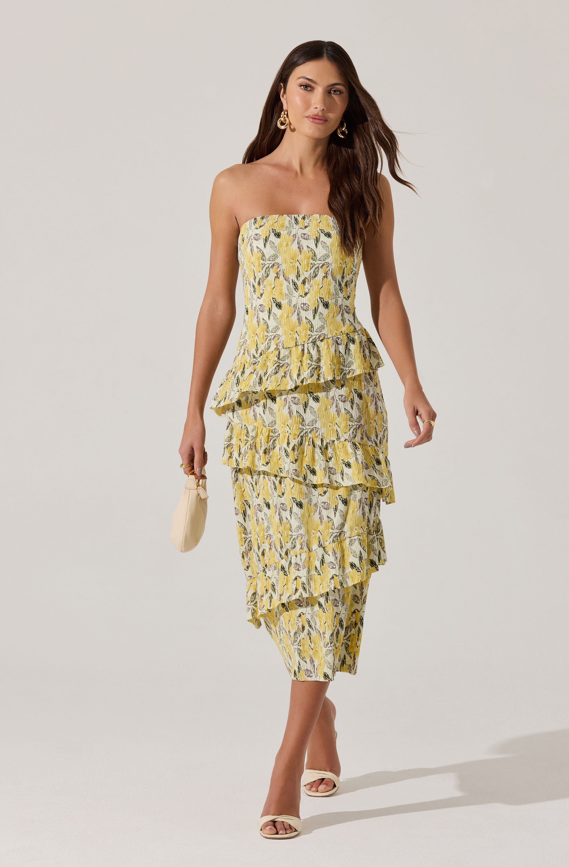 A woman is wearing a strapless, yellow and gray floral patterned dress with a layered, ruffled design. She accessorizes with large, gold hoop earrings, a small, light-colored clutch, and open-toe heels.