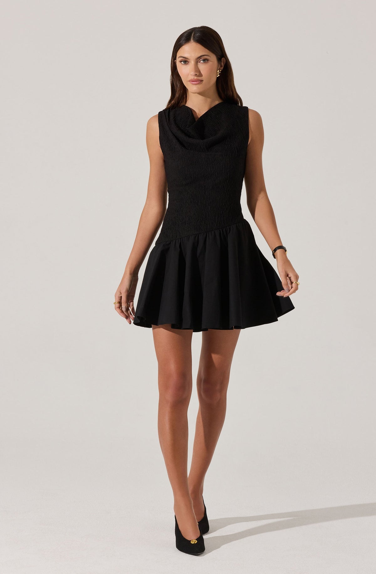 A woman is wearing a sleeveless black dress with a fitted, textured bodice and a wide, gathered skirt that flares out above the knees. The dress has a high neckline with a draped detail. She is also wearing black high-heeled shoes.