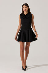 A woman is wearing a sleeveless black dress with a fitted, textured bodice and a wide, gathered skirt that flares out above the knees. The dress has a high neckline with a draped detail. She is also wearing black high-heeled shoes.