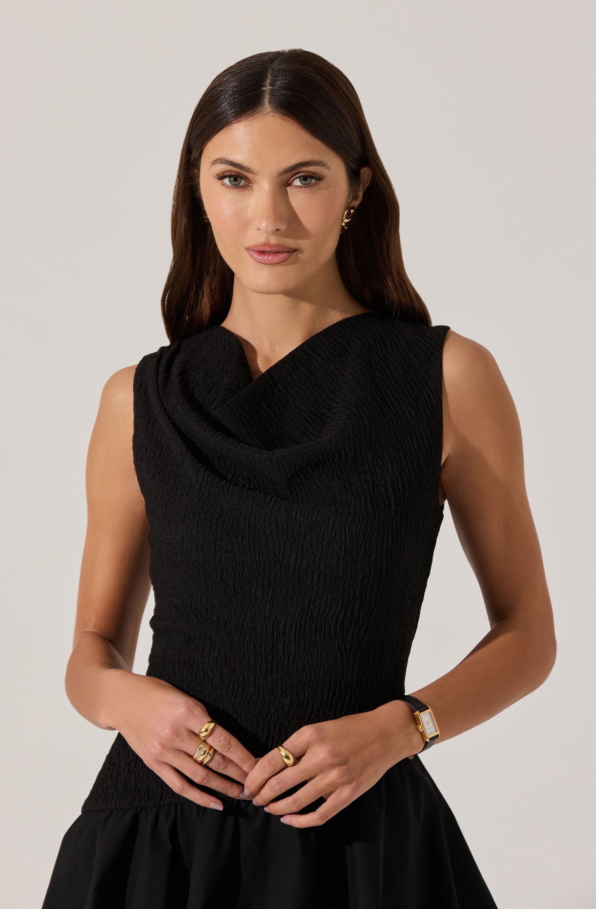 A woman is wearing a sleeveless black top with a textured, ruched fabric and a draped cowl neckline. She is accessorized with gold earrings, multiple rings, and a gold wristwatch.
