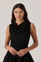 A woman is wearing a sleeveless black top with a textured, ruched fabric and a draped cowl neckline. She is accessorized with gold earrings, multiple rings, and a gold wristwatch.