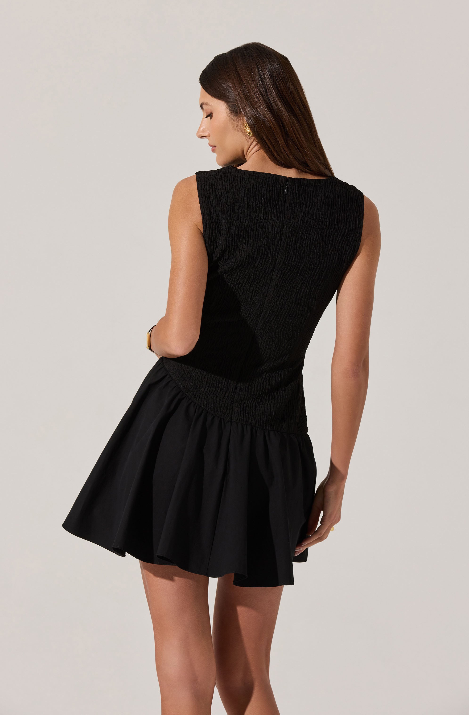 A woman is wearing a sleeveless black dress with a textured, fitted bodice and a flared, gathered skirt. The dress appears to have a high neckline and is made of a fabric with a subtle sheen.