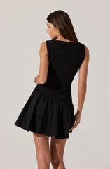 A woman is wearing a sleeveless black dress with a textured, fitted bodice and a flared, gathered skirt. The dress appears to have a high neckline and is made of a fabric with a subtle sheen.