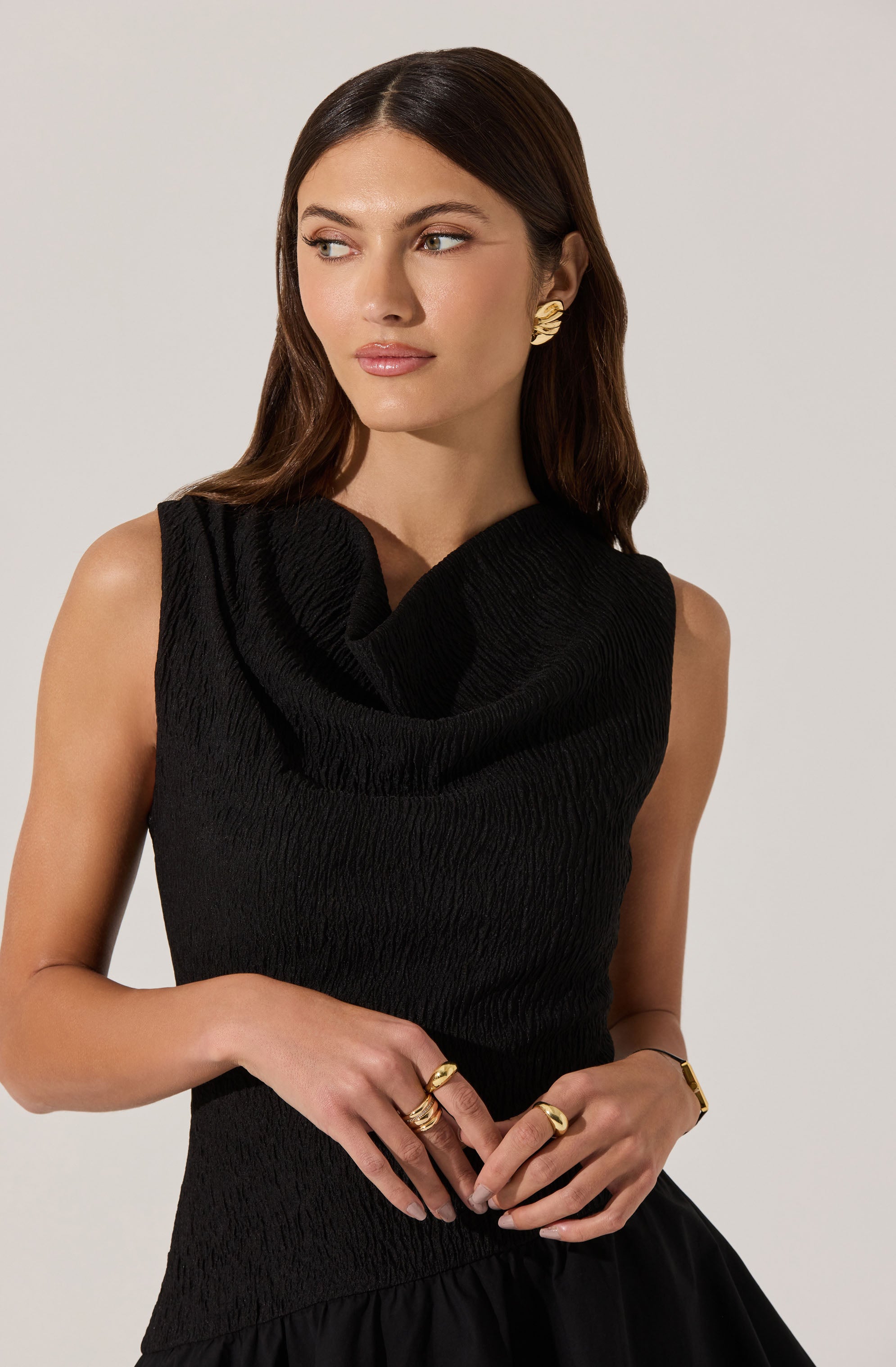 A woman is wearing a sleeveless black dress with a textured, slightly crinkled fabric. The dress has a wide, draped collar that folds over at the neckline. She accessorizes with large gold leaf-shaped earrings and multiple gold rings on her fingers.