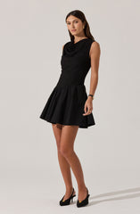 A woman is wearing a sleeveless black dress with a textured, ruched bodice that has a loose, draped neckline. The skirt is short, flared, and has a smooth fabric with a slightly shiny finish. She is accessorized with gold earrings, a black bracelet, and gold ring, and completes her outfit with black high-heeled shoes featuring small gold accents on the toes.