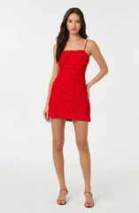 A woman is wearing a sleeveless, red dress with thin spaghetti straps. The dress has a textured, floral pattern throughout and is fitted at the waist, ending above the knees. She is also wearing red, open-toe high-heeled sandals.
