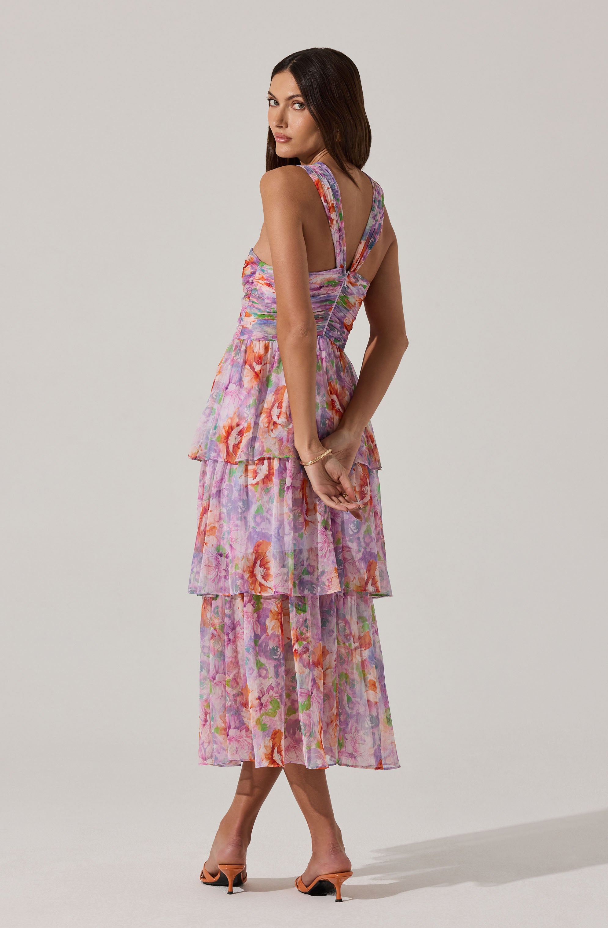 A woman is wearing a sleeveless, tiered dress with a floral pattern in shades of pink, purple, and orange. The dress has wide straps and a fitted bodice with a slight V-neckline. The skirt consists of three layers of flowing, semi-sheer fabric with a colorful floral design. She is also wearing open-toe high heel shoes and a delicate bracelet on her left wrist.