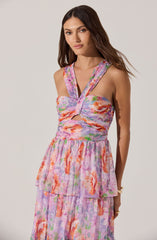 The woman is wearing a sleeveless, floral dress with a fitted bodice featuring a twisted front design and cut-out detail at the midriff. The dress has wide shoulder straps and a layered, flowing skirt with a pastel-colored floral pattern in shades of pink, orange, purple, and green.