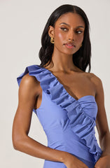 A woman is wearing a light blue, sleeveless dress featuring a large ruffle detail that runs from the shoulder down the front of the dress, creating a diagonal line across the chest. The dress has a fitted bodice and a sweetheart neckline. She is accessorized with gold hoop earrings.