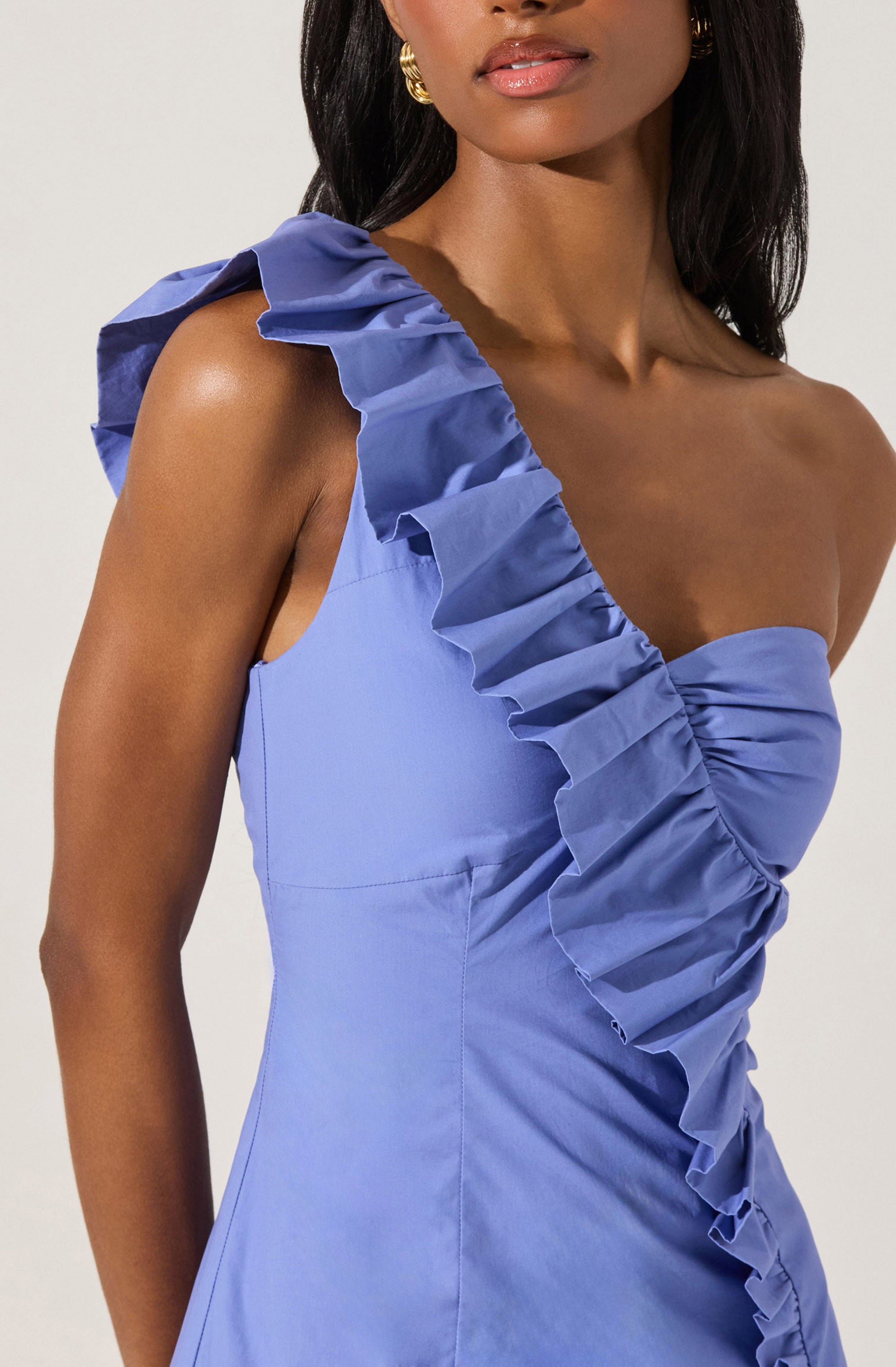 A woman is wearing a light blue, one-shoulder dress with ruffled detailing along the strap and bodice. The dress features an asymmetrical design with a prominent ruffle that extends from the shoulder across the chest. The fabric appears structured and smooth. She accessorizes with gold earrings and has dark, wavy hair.