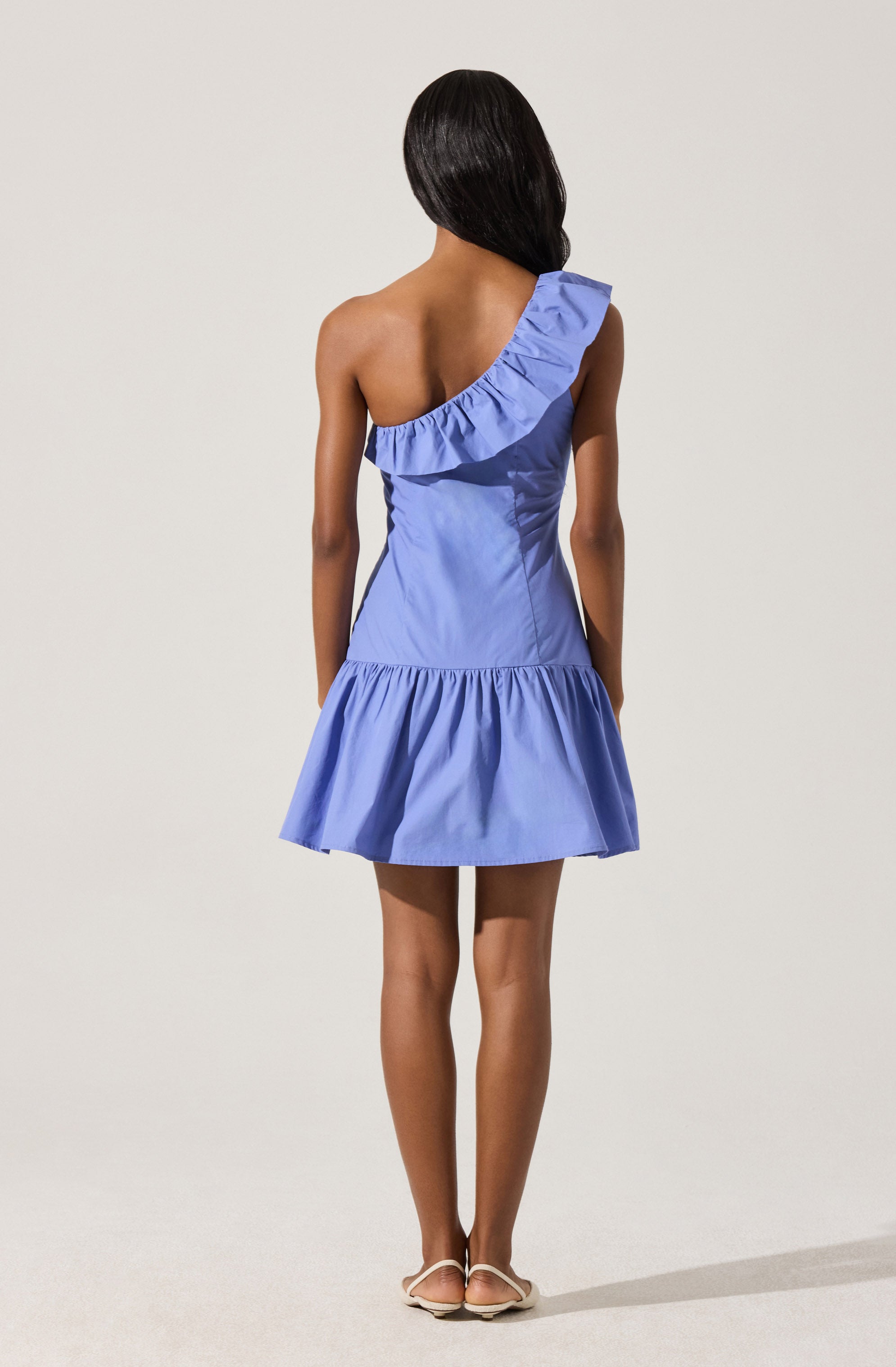 A woman is wearing a light blue, sleeveless dress with a ruffled one-shoulder neckline and an A-line skirt that flares out at the hem. She is standing against a plain light background, with her back facing the camera. The dress features subtle vertical seam details and gathered fabric at the waist.