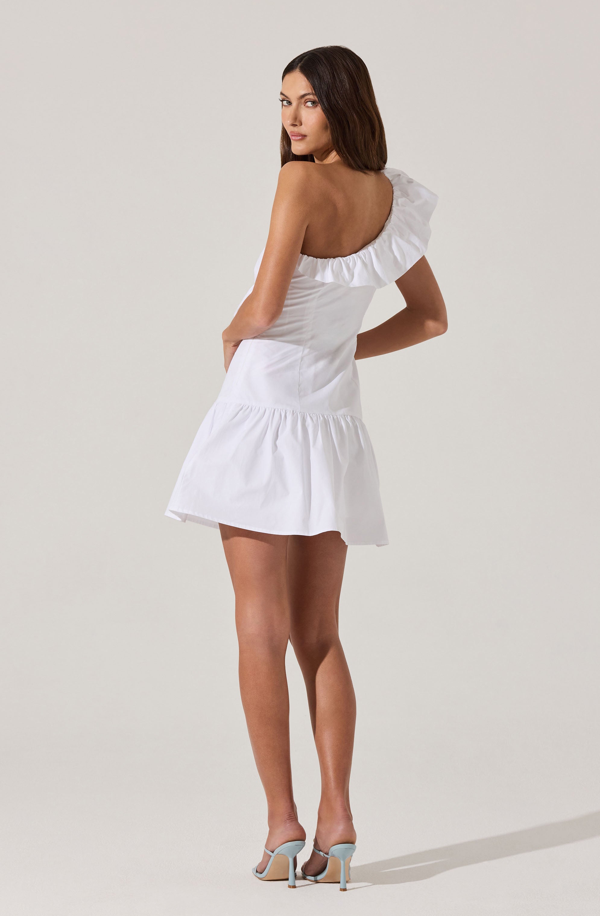 A woman is wearing a white, one-shoulder dress with a ruffled neckline and a tiered skirt. She is also wearing high-heeled shoes that are light-colored with a strap around the ankle.
