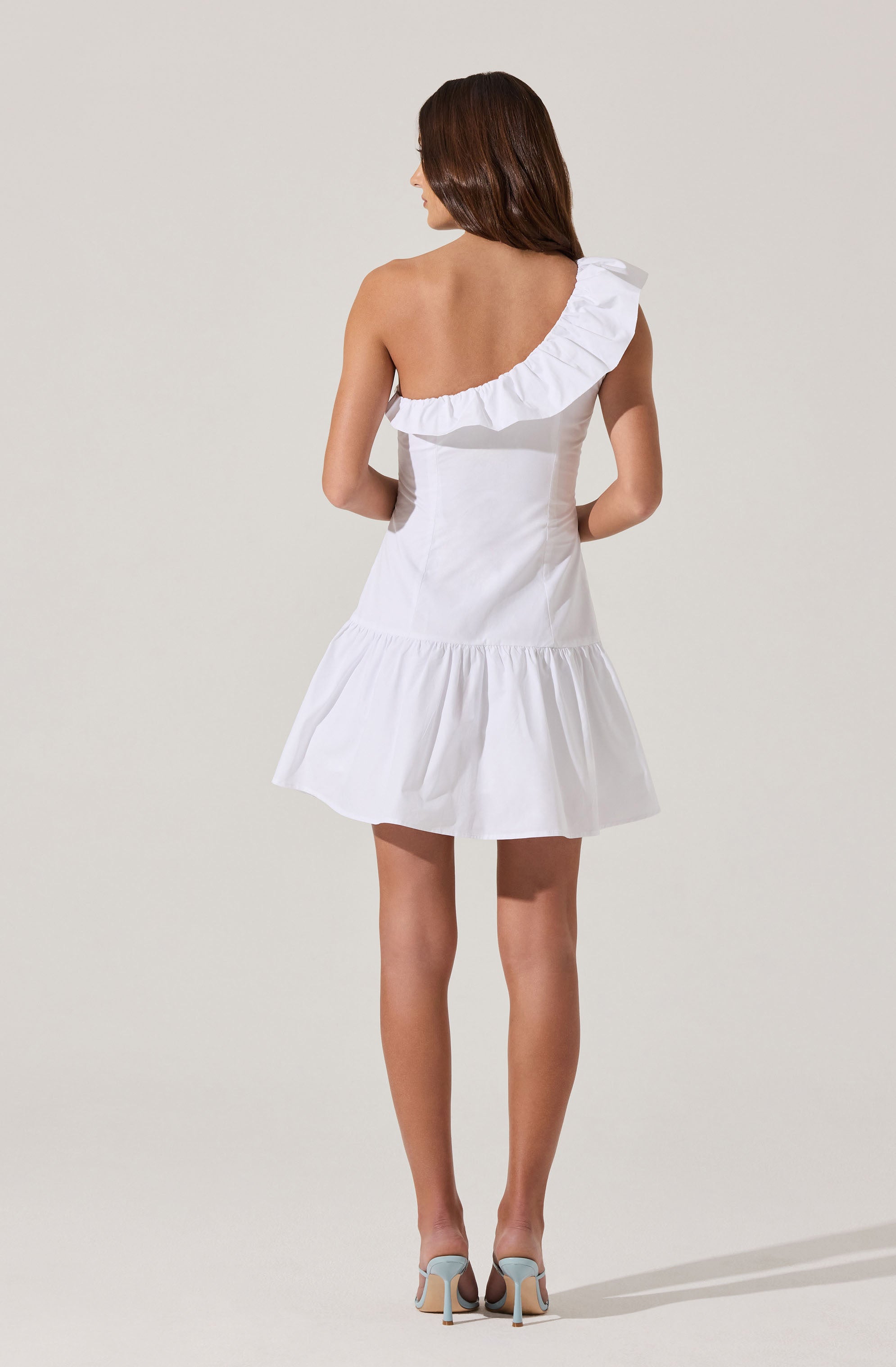 A woman is wearing a white, off-shoulder, sleeveless dress with ruffle details along the neckline and a gathered hemline that falls above the knees. She is also wearing light-colored high-heeled shoes.