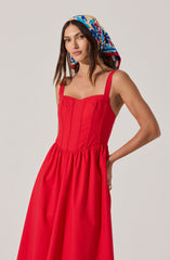 A woman is wearing a sleeveless, bright red dress with a fitted bodice that features vertical seams and a sweetheart neckline. The skirt is full and gathered at the waist. She has a colorful scarf with a floral pattern in shades of blue, red, beige, and white wrapped around her head.