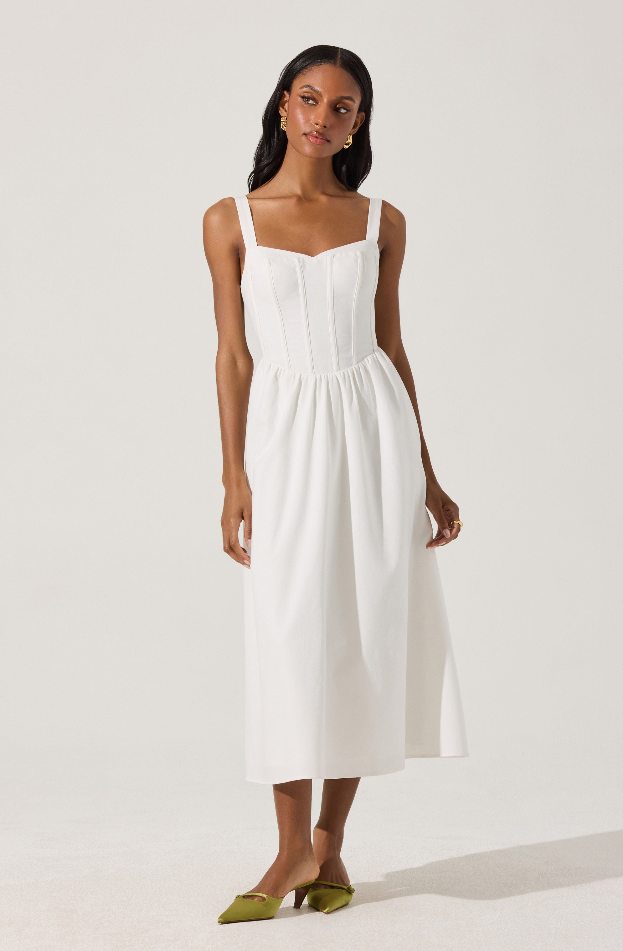 A woman is wearing a white sleeveless dress with wide straps and a fitted bodice that features vertical seams. The dress has a gathered waistline and a long, flowing skirt that reaches mid-calf. She is also wearing a pair of green pointed-toe heels with a small heel.