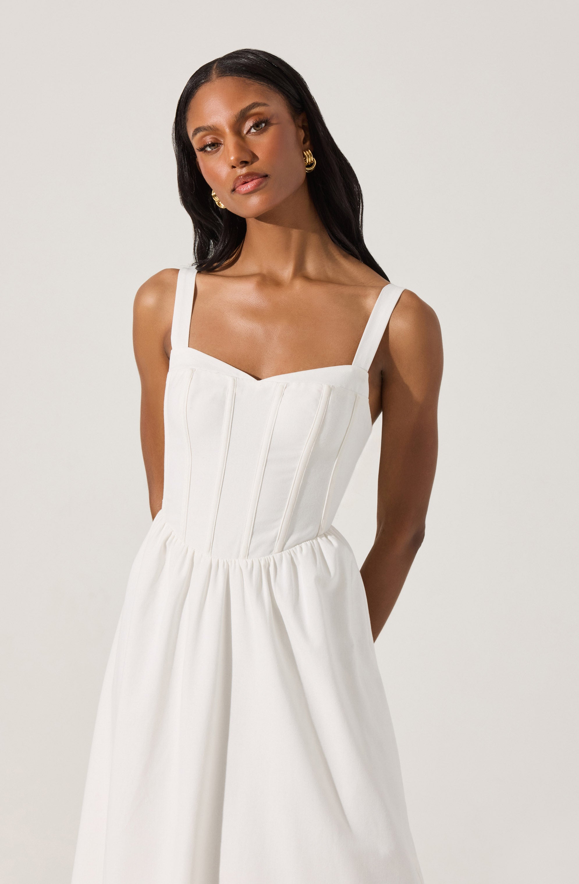 A woman is wearing a sleeveless white dress with wide shoulder straps. The dress features a fitted bodice with vertical seam details and a gathered waistline that creates a flowing skirt.