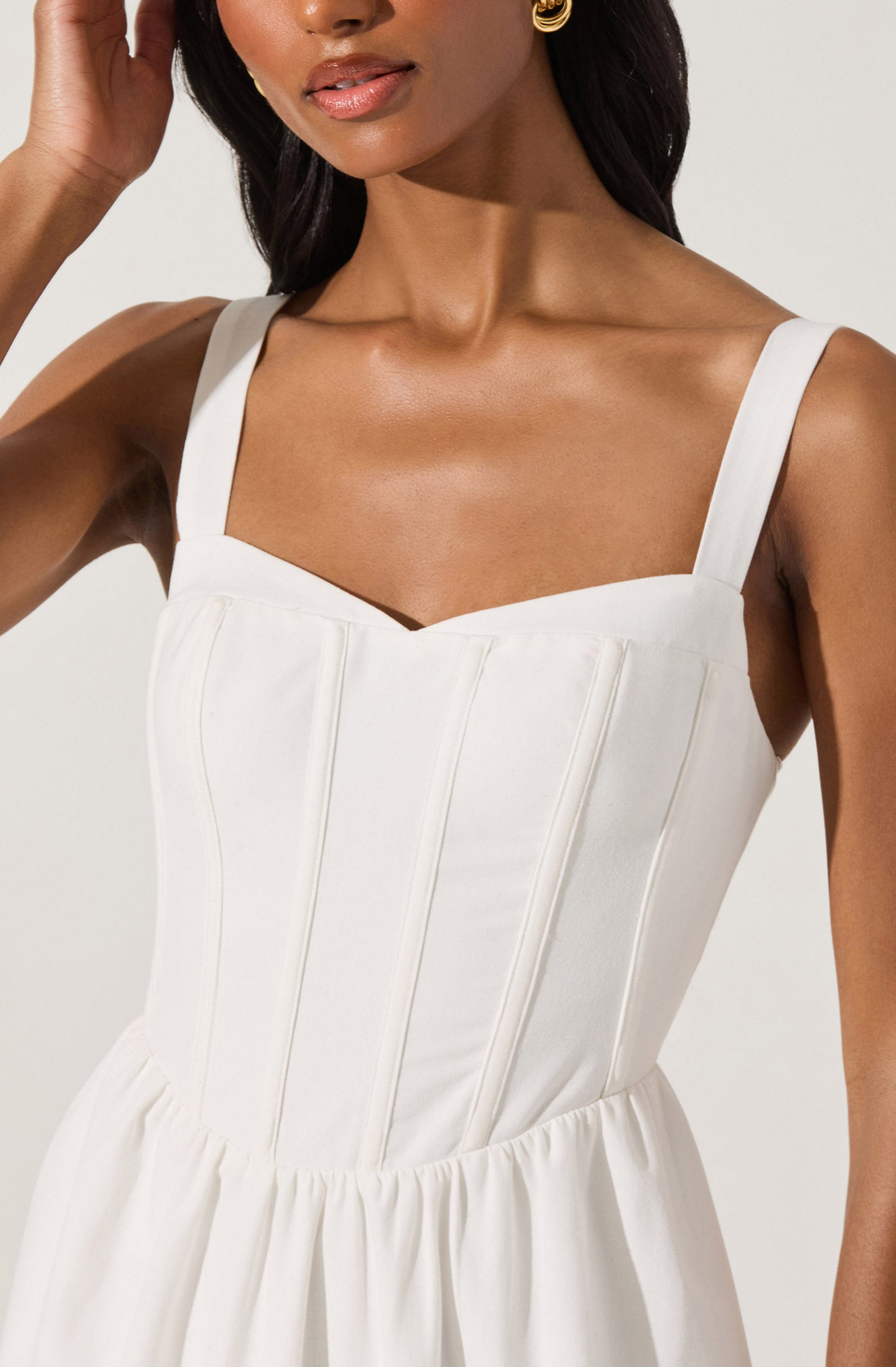 A woman is wearing a sleeveless white dress with a fitted bodice featuring vertical seams and wide shoulder straps. The dress has a gathered waistline and a flowing skirt. She has dark hair and is accessorizing with gold hoop earrings.