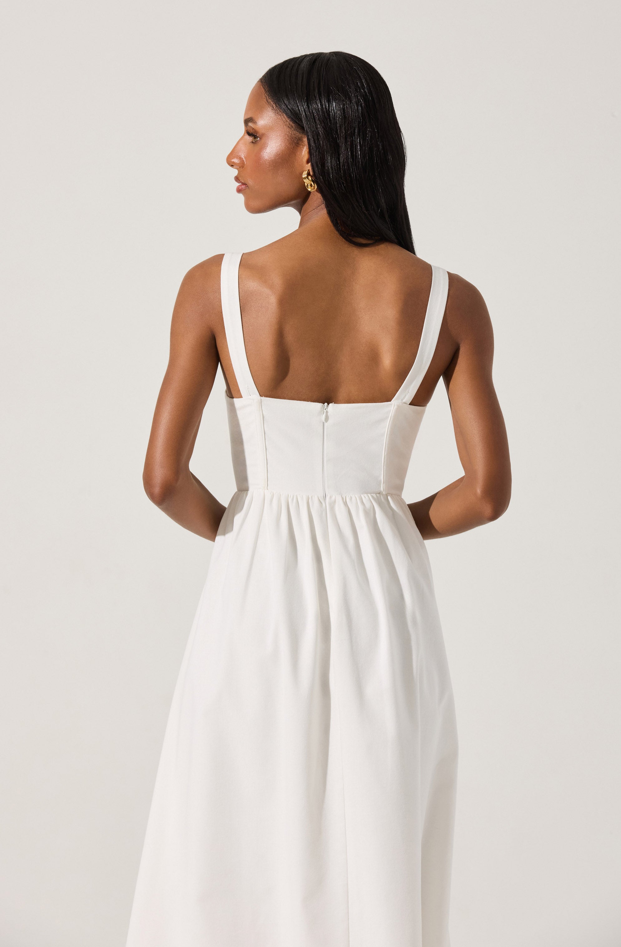 A woman is wearing a sleeveless, white dress with wide shoulder straps, a fitted bodice, and a high waistline. The dress has a back zipper closure and a flowing skirt. She has dark, straight hair and is accessorized with gold earrings.