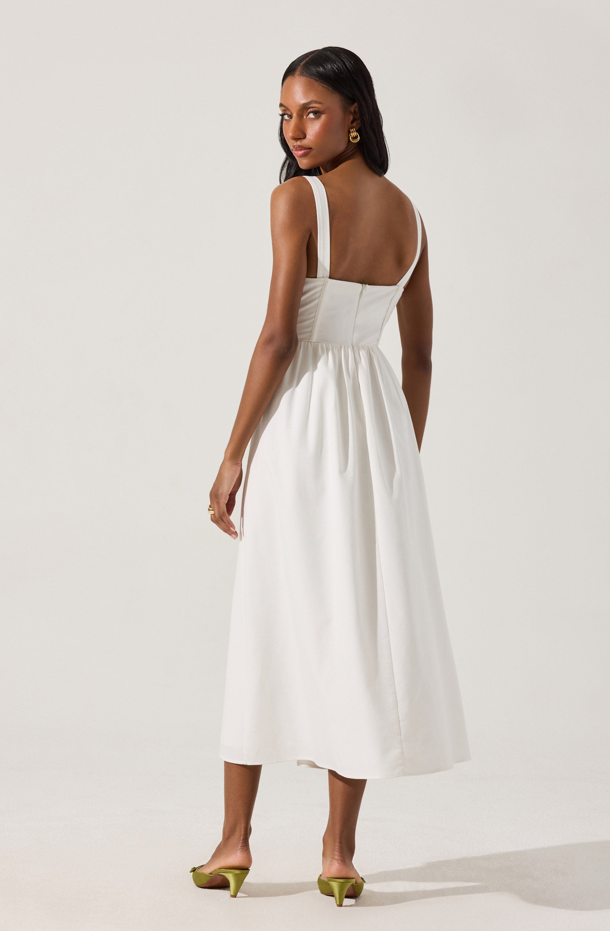 A woman is wearing a sleeveless white dress with wide shoulder straps, a fitted bodice, and a gathered skirt that falls to mid-calf length. She has gold hoop earrings and stands against a plain, light-colored background.