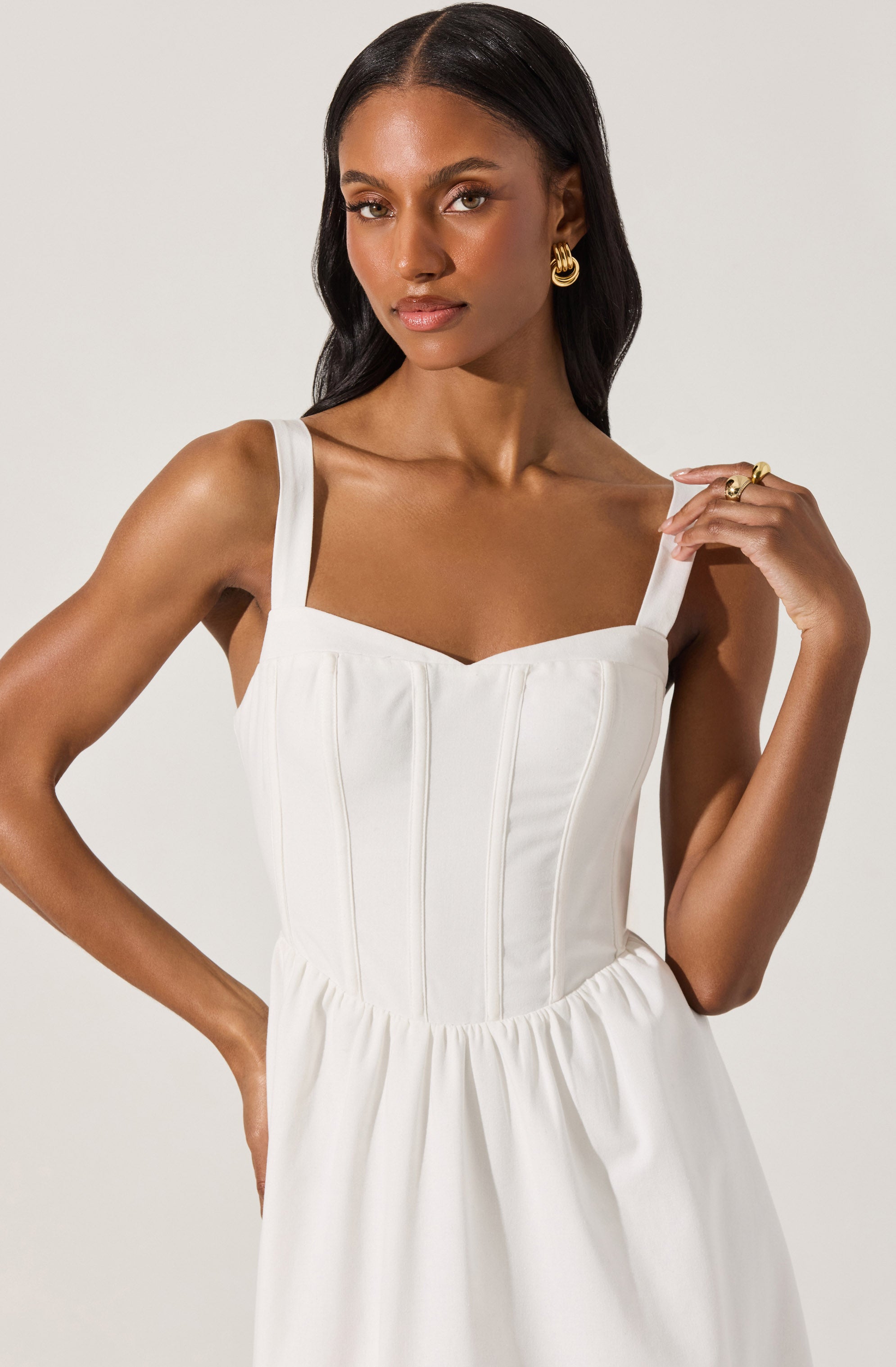 A woman is wearing a white dress with wide shoulder straps and a fitted bodice featuring vertical seams. The dress has a gathered waistline, creating a slightly billowy skirt. She accessorizes with gold hoop earrings and rings, and has long dark hair styled in soft waves.