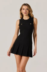 A woman is wearing a sleeveless black dress with a round neckline. The dress features two small decorative pockets with gold buttons on the chest and a pleated skirt that falls above the knees.