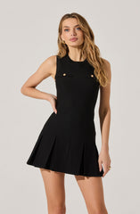 A woman is wearing a sleeveless black dress with a fitted bodice and a pleated skirt. The top of the dress features two small decorative gold buttons on the chest and a small horizontal black strap detail. The skirt has evenly spaced pleats, giving it a structured and elegant appearance.