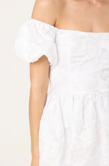 A white dress with a floral textured pattern, featuring puffed short sleeves with an elasticated ruffle at the shoulder. The fabric appears to be lightweight and embroidered with large, overlapping flower motifs.