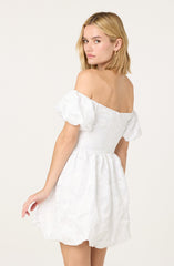 A woman is wearing an off-the-shoulder white dress with puffed short sleeves, a fitted bodice, and a slightly flared skirt. The dress has a textured fabric with a subtle floral or embroidered pattern.
