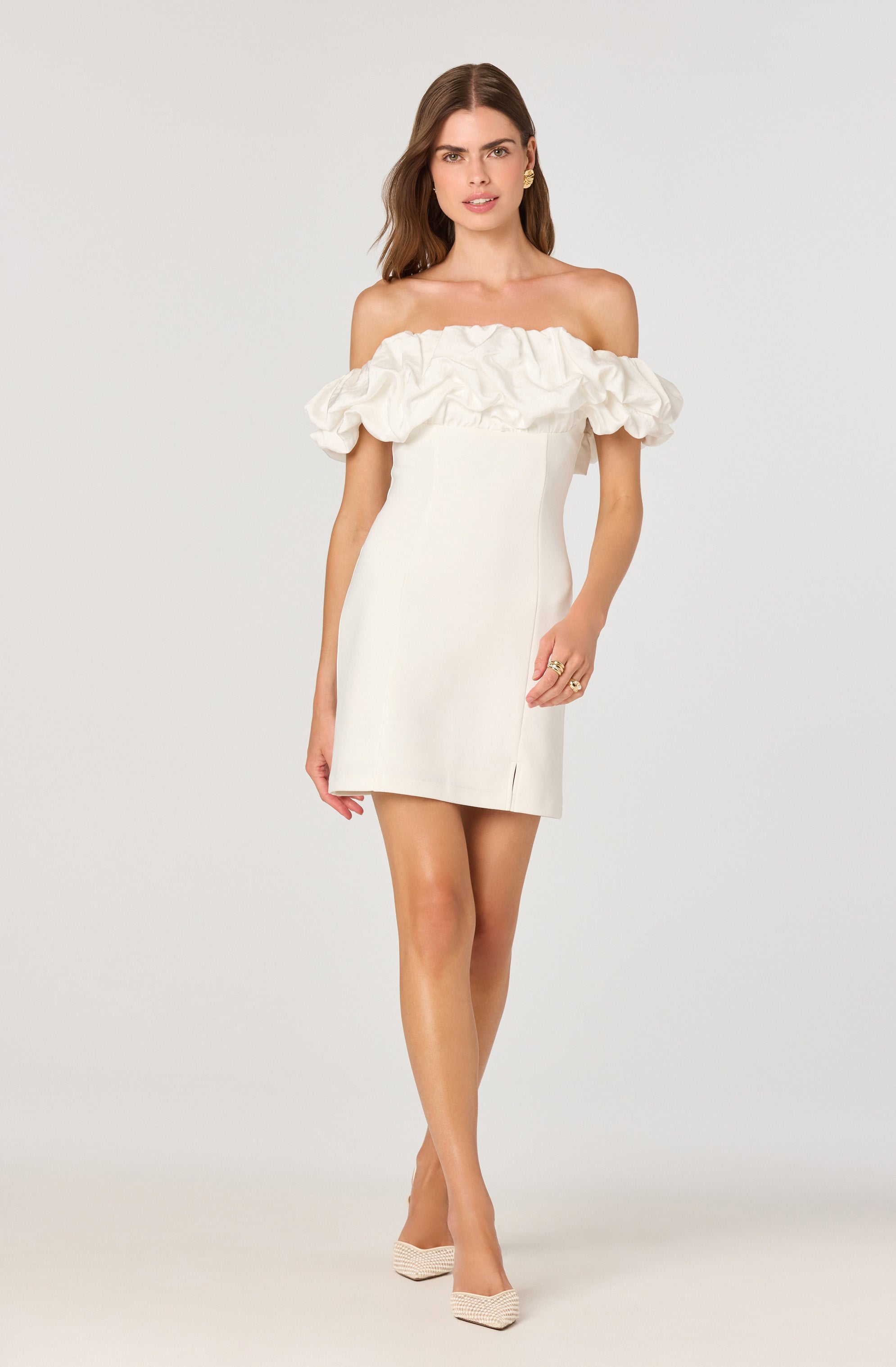 A woman is wearing an off-the-shoulder white dress with a ruffled neckline detail. The dress is fitted and short in length, ending mid-thigh. She is also wearing beige high-heeled shoes.