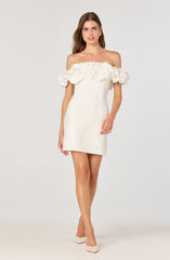 A woman is wearing an off-the-shoulder white dress with a ruffled neckline detail. The dress is fitted and short in length, ending mid-thigh. She is also wearing beige high-heeled shoes.