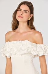 A woman is wearing an off-the-shoulder white dress with a large, ruffled, and gathered fabric detail around the neckline. She has on bold gold earrings and has shoulder-length brown hair.