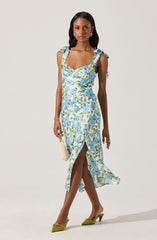 The woman is wearing a sleeveless, ankle-length dress with a light-colored background and a floral pattern featuring blue, white, and green colors. The dress has shoulder straps tied into bows. She is also wearing green pointed-toe high heels and carrying a small, white handbag.