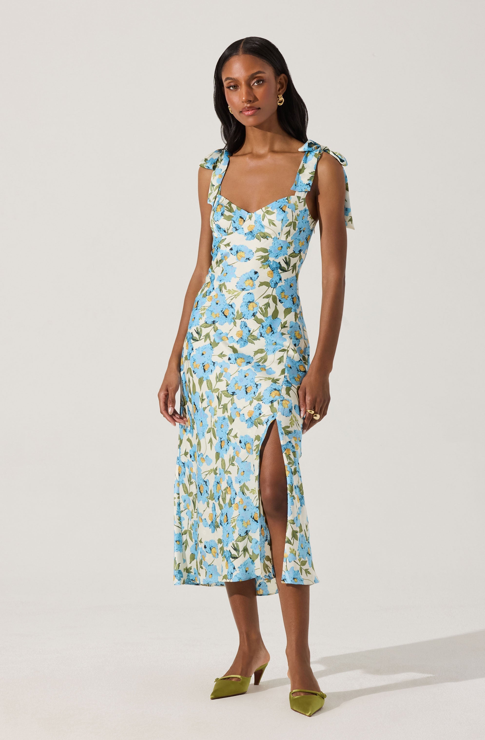 A woman is wearing a sleeveless, ankle-length dress with a floral pattern featuring blue, white, and green colors. The dress has shoulder straps tied into bows and a slit on the front left side. She is also wearing green pointed-toe heels.