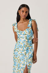 A woman is wearing a sleeveless dress with shoulder ties that form bows. The dress features a floral pattern with blue flowers and green leaves on a light background. It has a sweetheart neckline and a slit on the right side. She accessorized with gold earrings and a ring.