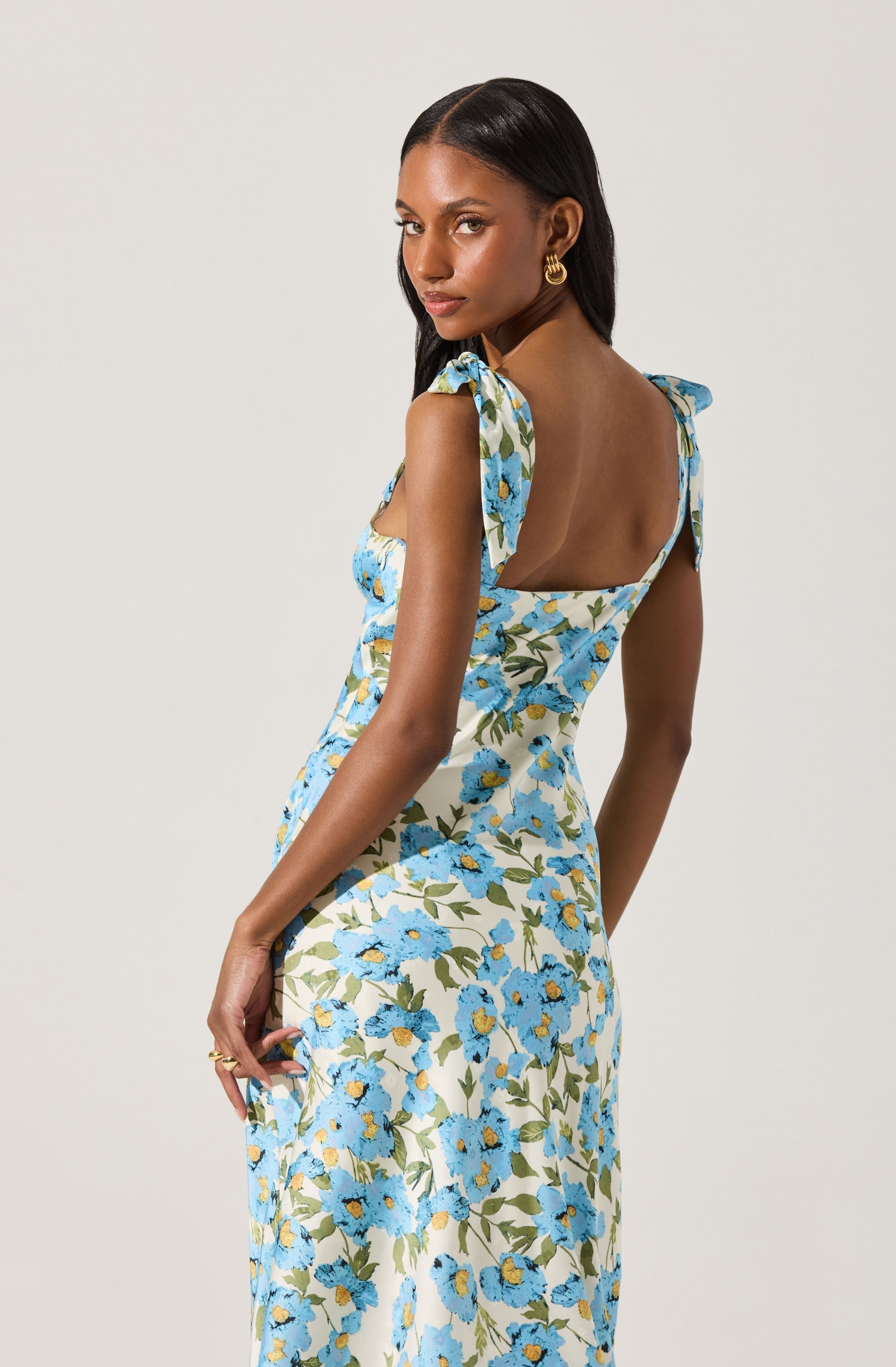 A woman is wearing a sleeveless, fitted dress with shoulder straps tied into bows. The dress features a floral pattern with blue flowers and green leaves on a light background. She is accessorized with large gold hoop earrings.