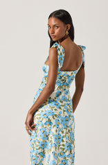 A woman is wearing a sleeveless, fitted dress with shoulder straps tied into bows. The dress features a floral pattern with blue flowers and green leaves on a light background. She is accessorized with large gold hoop earrings.