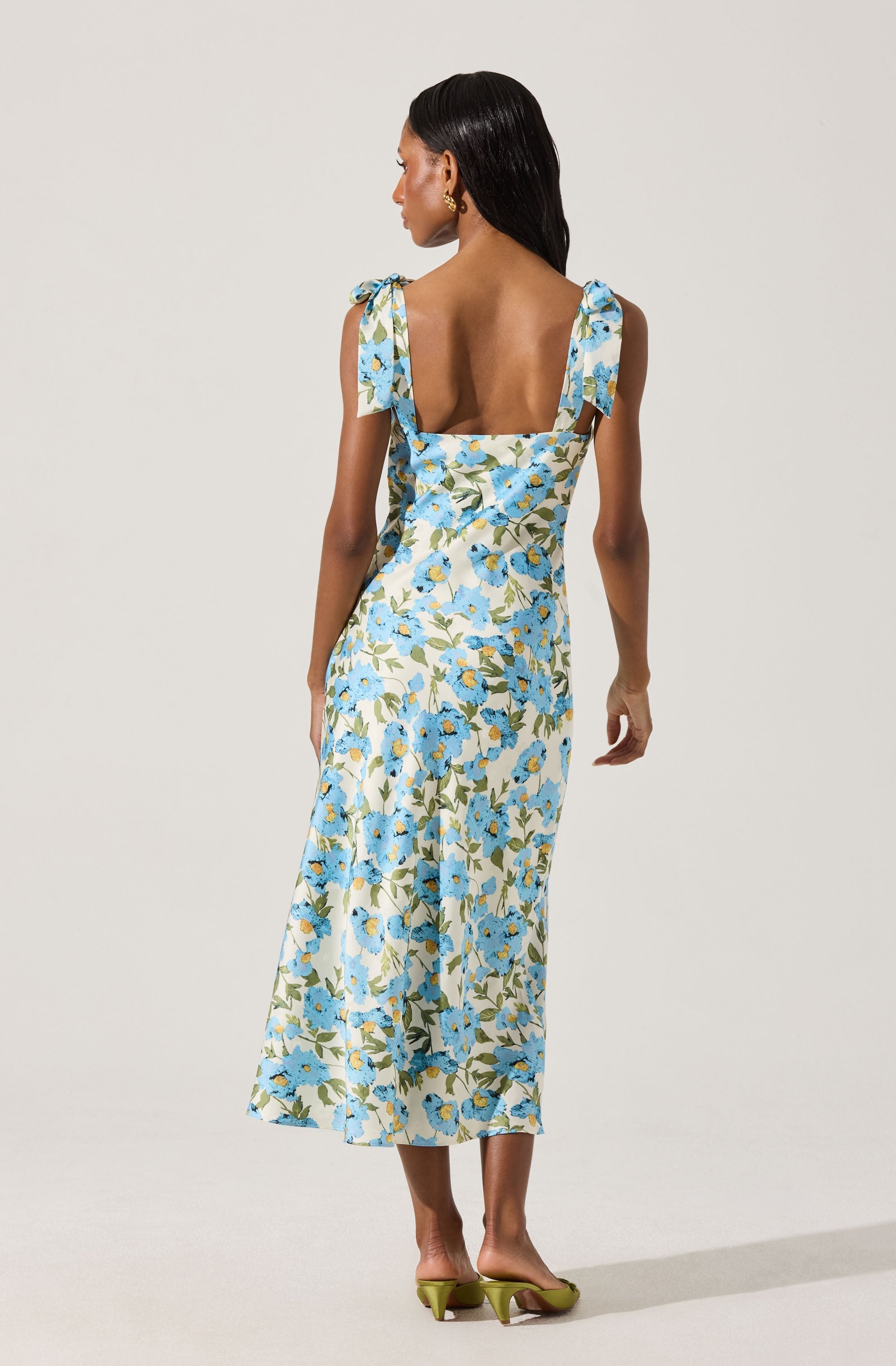 A woman is wearing a long, sleeveless dress with shoulder tie straps. The dress features a light background with a blue floral pattern and green leaves. She is wearing green high-heeled shoes.