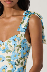 A woman is wearing a sleeveless dress with a floral pattern featuring blue flowers with yellow centers and green leaves on a white background. The dress has a shoulder tie detail with a bow made from the same fabric.