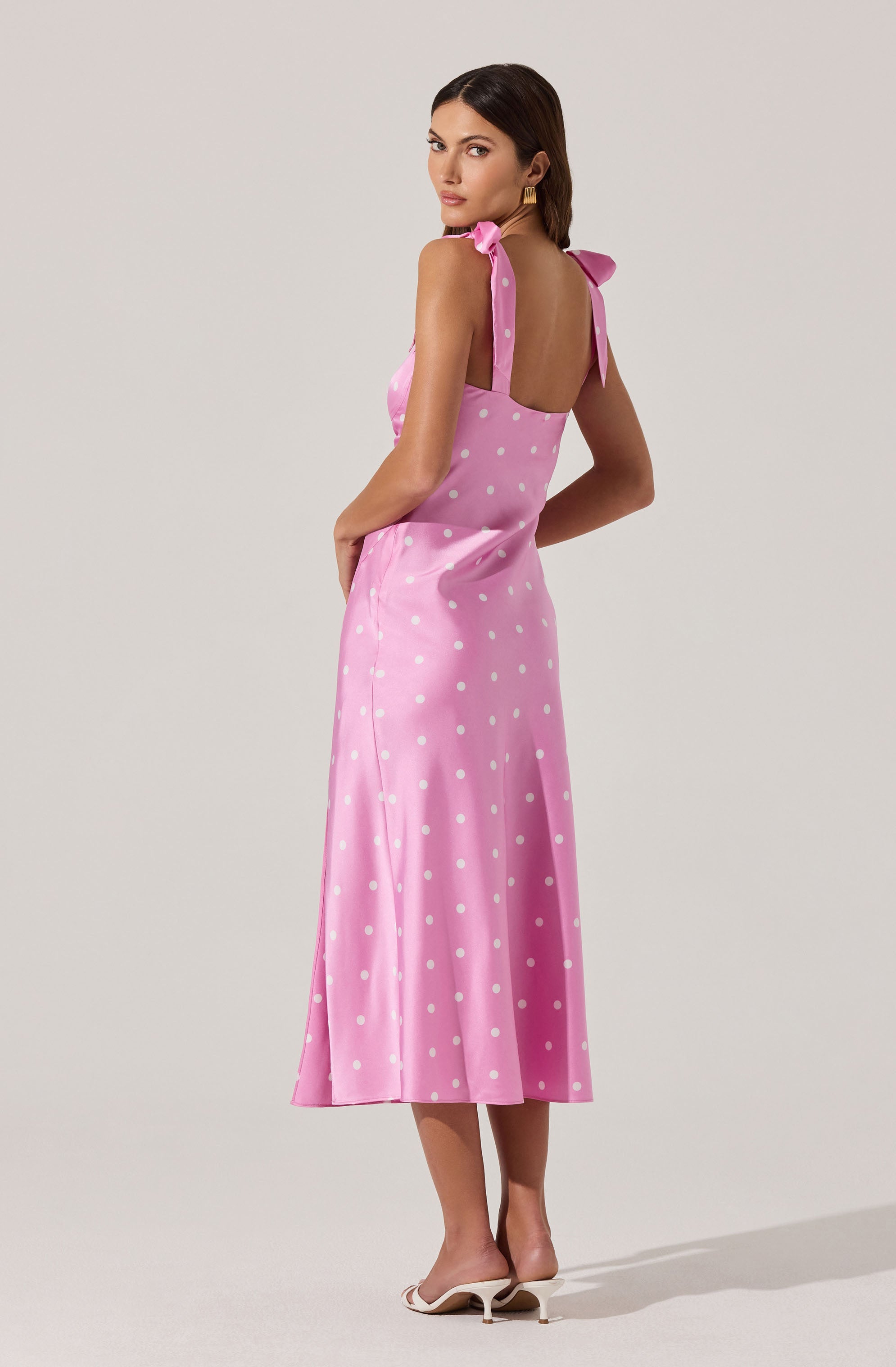 A woman is wearing a pink sleeveless midi dress with white polka dots. The dress features shoulder straps tied into bows on each shoulder and a fitted bodice that flows into a slightly flared skirt. She is also wearing white heeled sandals.