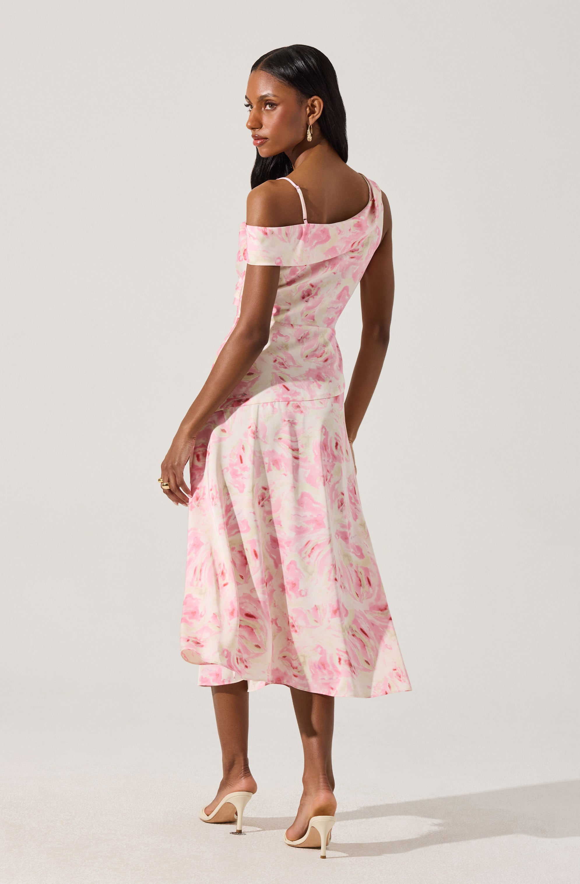 A woman is wearing an off-the-shoulder dress with spaghetti straps. The dress has a pink and white marbled pattern, fitting snugly at the top and flowing into a midi-length skirt. She is also wearing nude high-heeled shoes.