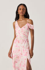 A woman is wearing a pastel pink and cream tie-dye dress with a V-neckline and thin spaghetti straps. The dress features a cold-shoulder ruffle detail on one side and a high slit on the leg.
