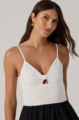 A woman is wearing a white sleeveless top with thin spaghetti straps. The top features a ribbed texture with a small central knot and a cut-out detail just below the knot. The top is fitted and extends to the waist, where it meets a black, gathered skirt. She has long, dark brown hair and is accessorized with gold square earrings.