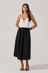 A woman is wearing a white sleeveless top with thin spaghetti straps and a small cutout detail at the front. She is also wearing a high-waisted, full-length black skirt with a gathered waistband. She has on black open-toe heels.