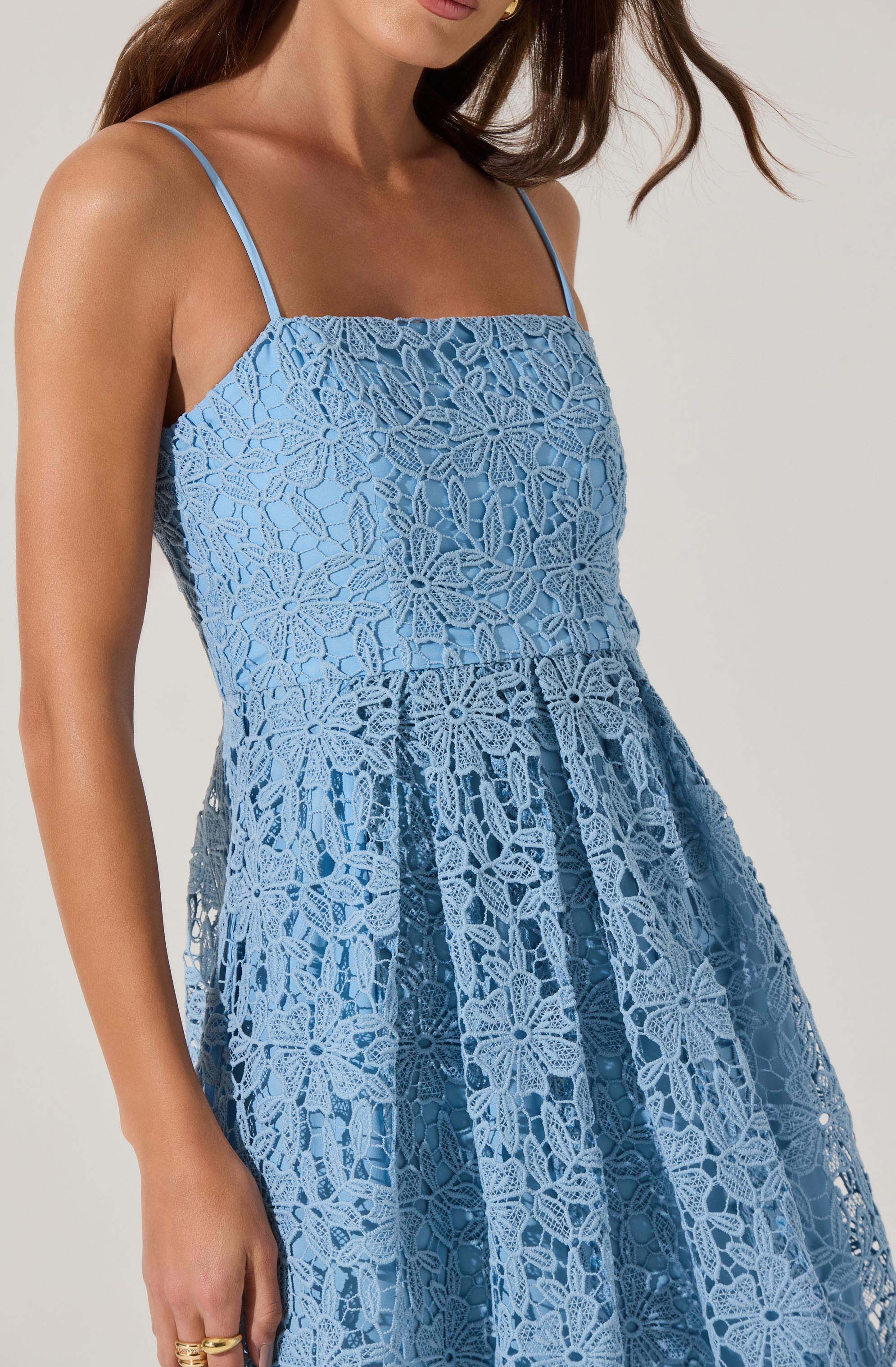 A woman is wearing a light blue dress with thin spaghetti straps. The dress features an intricate lace overlay with floral and geometric patterns, creating a textured and delicate appearance. The skirt of the dress has pleats, adding volume and movement.